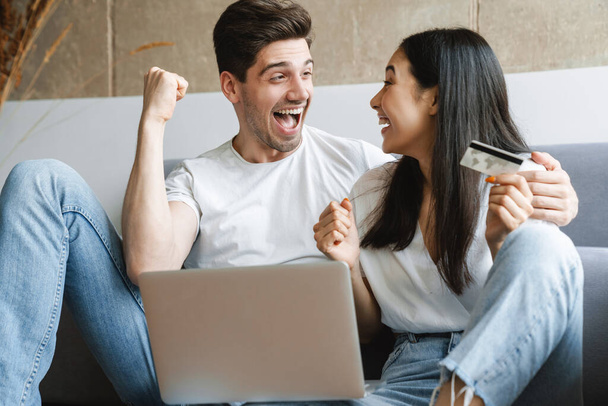 Image of excited cheery young loving couple at home on floor near sofa using laptop computer and making a winner gesture - Foto, immagini
