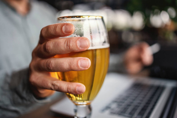 Closeup of man holding a glass of fresh cold light beer and working on laptop. - Photo, Image