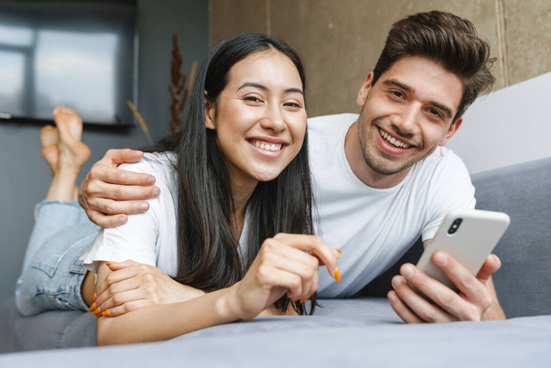 Happy couple enjoying media content in a smart phone while relaxing on a couch at home - Fotoğraf, Görsel