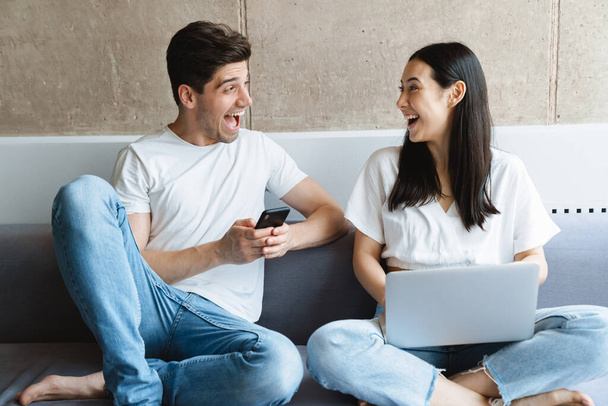 Image of a cheerful young loving couple at home on sofa using laptop computer and mobile phone - Valokuva, kuva
