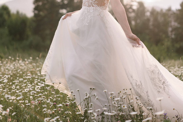 Nevěsta ve svatebních šatech při západu slunce kráčí po heřmánkovém poli - Fotografie, Obrázek