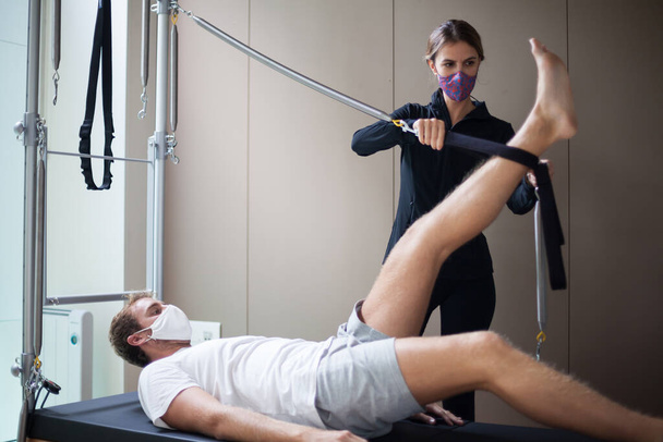 joven hombre usando máscara haciendo ejercicios de pilates en reformador con su instructor femenino durante covid-19 - Foto, imagen