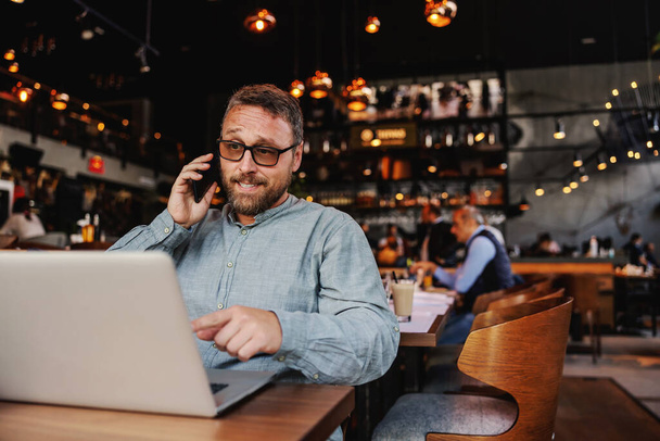 Propietario de café barbudo de mediana edad con anteojos sentados en un bar, hablando con un asesor financiero y apuntando a la computadora portátil. - Foto, imagen