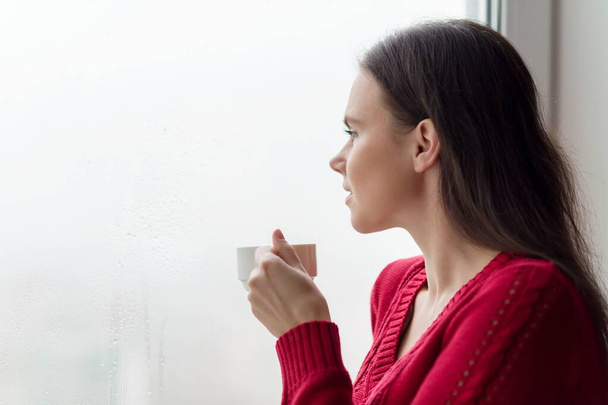 Young smiling woman enjoying coffee, female in warm knitted sweater near the window - Foto, Bild