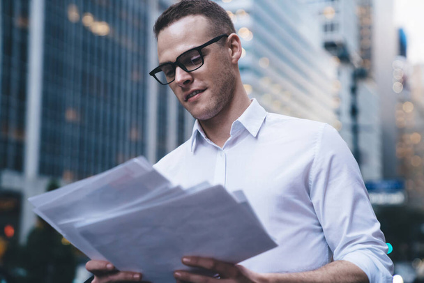 Young good looking executive in casual clothes and fashionable glasses reviewing information on paper while standing on blurred background of modern skyscrapers - Фото, зображення