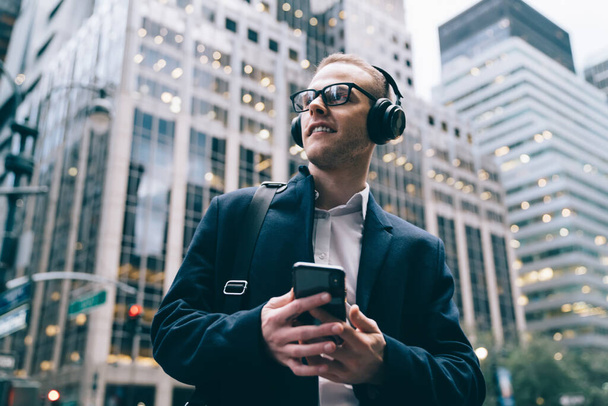 From below young male entrepreneur in formal attire and glasses standing in downtown while listeinging to music from smartphone through headphones - 写真・画像