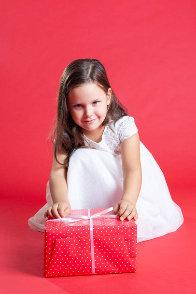 smiling child in a white dress sitting on his haunches and unleashes a red box, the receipt of gifts - Foto, Bild