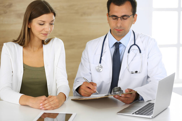 Doctor and patient discussing the results of a physical examination while sitting at a desk in a clinic. A male doctor using a clipboard to fill out a medical history of a young womans medication - Zdjęcie, obraz