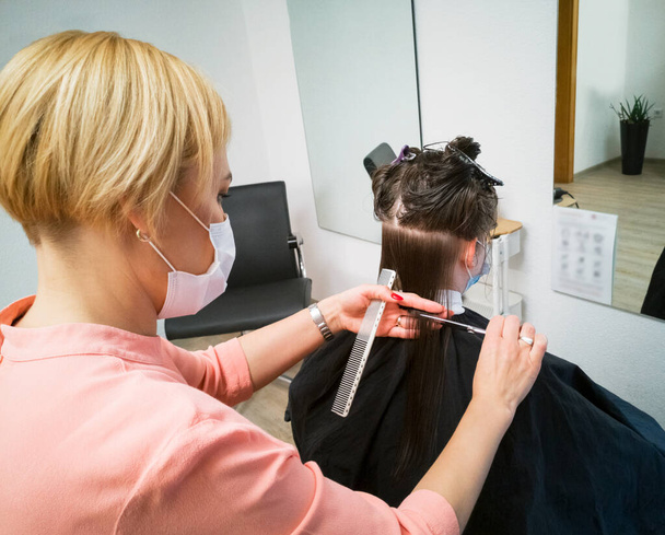 Siyah-19 güvenlik önlemleri için tıbbi maskeli sarışın kuaför esmer kadın müşteri saçını kesti. Güzellik salonunun yeniden açılması, pandemik karantina. Klasik kuaför teknolojisi.. - Fotoğraf, Görsel