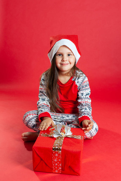 a girl in pajamas and a Santa Claus hat is sitting barefoot on the floor next to a Giftbox on a red background, isolated - Photo, Image