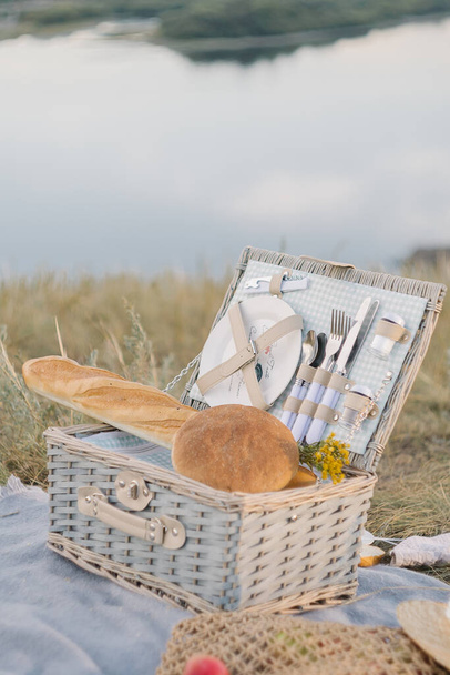 Picnic set with dishes, bread, fruits and hat on the river bank - Fotoğraf, Görsel