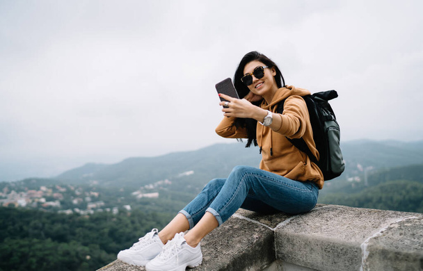 Cheerful asian girl in sunglasses resting during vacation trip visiting historical place making picture on smartphone, happy female tourist posing for selfie using mobile phone camera on destination - 写真・画像