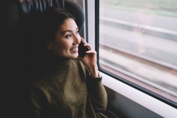 Hymyilevä nainen lämpimässä puserossa istuu ikkunapenkillä ja katselee näkymää ulkona rentoutuen junassa ja vastaten puheluun - Valokuva, kuva