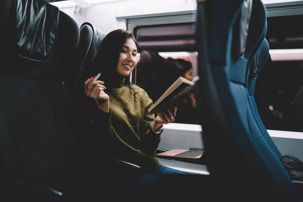 Smiling asian hipster girl reading book during trip in comfortable train sitting in seat holding literature, cheerful female blogger reading article for publication satisfied with trip and train for remote job - Fotoğraf, Görsel
