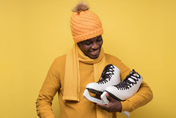African young man in yellow hat and scarf with winter skates on yellow background. - Photo, Image