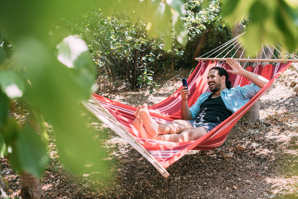 Молодой человек отдыхает и разговаривает по телефону в гамаке в саду. Красивый парень лежит расслабленный в гамаке и общается на смартфоне в тени деревьев в летний день - Фото, изображение