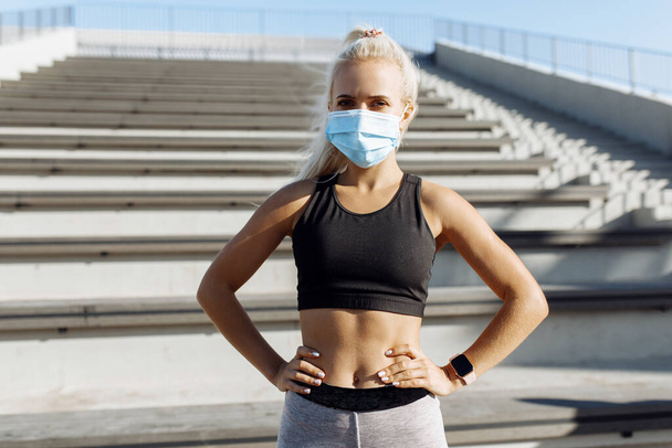 sportswoman in a medical protective mask on her face and sportswear stands in the city against the background of the stairs, the concept of a healthy lifestyle, quarantine, coronavirus - Foto, Bild