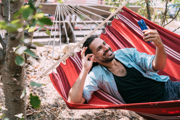 Un joven está descansando y hablando por teléfono en una hamaca en el jardín. Chico guapo se encuentra relajado en una hamaca y se comunica en un teléfono inteligente a la sombra de los árboles en un día de verano - Foto, Imagen