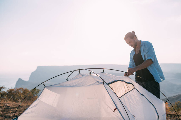 若い男性の観光客は山の中にテントを置く。男はハイキングにキャンプを設置し、日没時に山の中でテントを集めて - 写真・画像