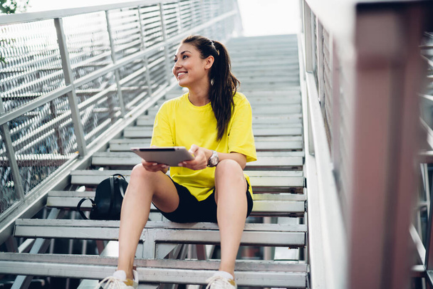 Smiling asian female in casual wear sitting on stairs urban setting holding digital tablet connected to 4G internet, positive hipster girl 20s millennial sending mails and messages on touchpad outdoors - Фото, изображение