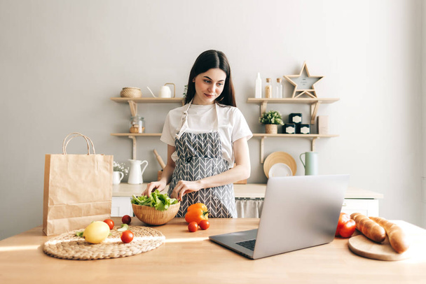 Young caucasian woman in apron use laptop computer in the modern kitchen, preparing salad, read recipe. - Fotoğraf, Görsel