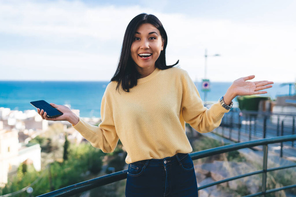 Half length portrait of joyful hipster girl with cellular technology in hand smiling at camera during travel sightseeing at overlooking area, cheerful female traveller shrugging in touristic town - Photo, image