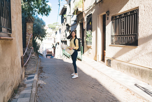 Full length portrait of cheerful Asian tourist smiling at camera during travel experience using paper location map for orientation navigation during solo vacations, happy hipster girl enjoying trip - Foto, immagini