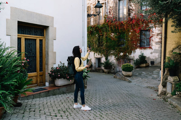 Side view of full length stylish woman in casual clothes and backpack standing near old building while walking along street - Fotografie, Obrázek