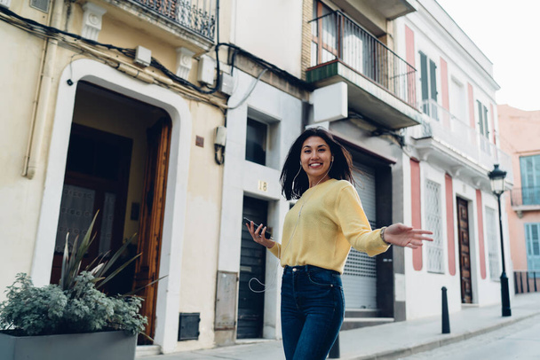 Half length portrait of cheerful tourist in headphones walking around touristic town and smiling at camera, joyful hipster girl with cellphone device using media app for listening music on leisure - Foto, Imagen