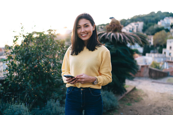 Cheerful Asian brunette female in casual clothes with toothy smile standing against bushes on street and browsing smartphone while looking at camera - Foto, Bild
