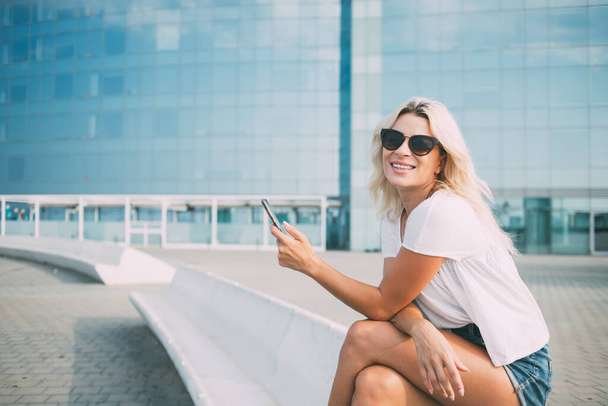 Portrait of cheerful hipster girl in stylish sunglasses holding modern cellular device and smiling at camera during leisure time in city, happy female user with smartphone posing at urban setting - 写真・画像