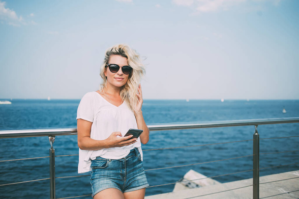 Half length portrait of millennial woman in trendy sunglasses looking at camera during time for summer promenade at seaside pier, Caucasian hipster girl holding modern smartphone technology - 写真・画像