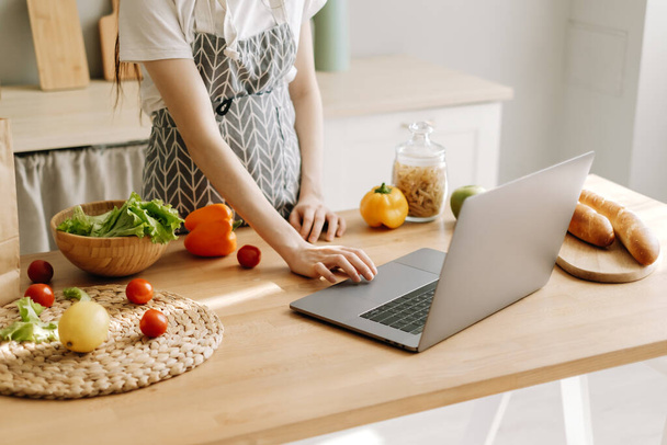 Young caucasian woman in apron use laptop computer in the modern kitchen, preparing salad, read recipe. - Φωτογραφία, εικόνα