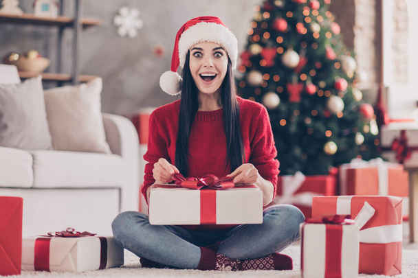 Photo of young lady sit floor hold unboxing gift open mouth wear santa cap red pullover jeans socks in decorated living room indoors - Foto, imagen