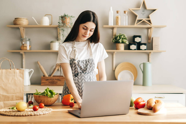 Young caucasian woman in apron use laptop computer in the modern kitchen, preparing salad, read recipe. - 写真・画像