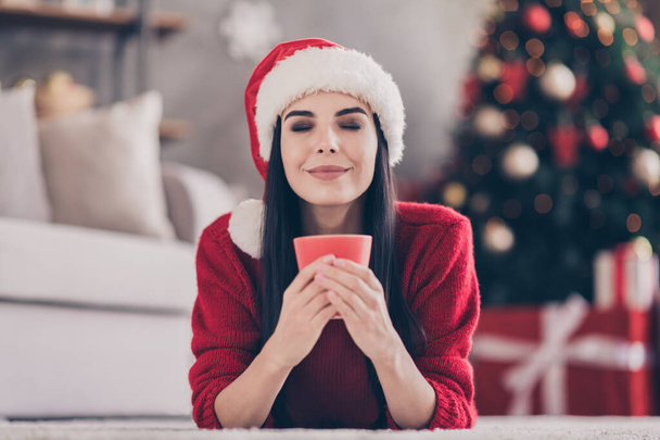 Photo of inspired cute girl lay floor hold cup closed eyes wear santa cap red pullover in decorated living room indoors - 写真・画像