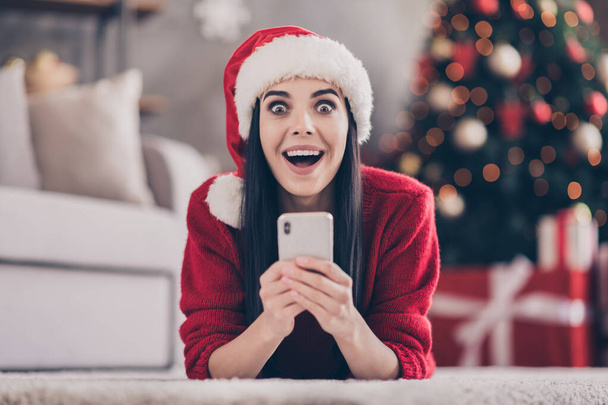 Photo of shocked lady lay floor hold smartphone open mouth wear santa cap red sweater in decorated living room indoors - Foto, Bild