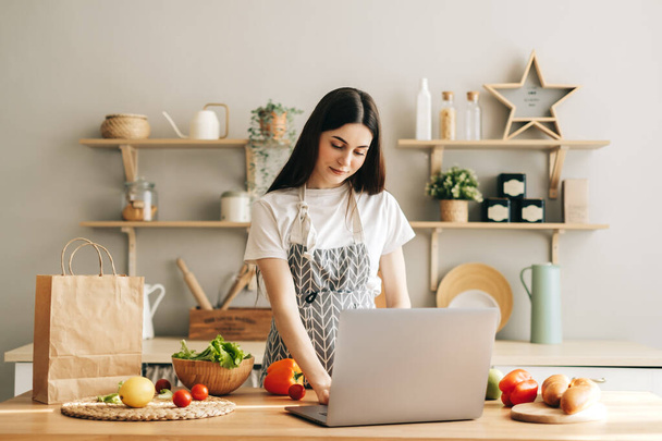 Young caucasian woman in apron use laptop computer in the modern kitchen, preparing salad, read recipe. - Zdjęcie, obraz