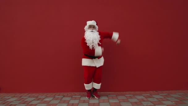 Lustiger Weihnachtsmann, der auf der Straße vor einer roten Wand tanzt und mit einem Lächeln im Gesicht in die Kamera schaut. - Filmmaterial, Video