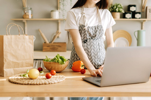 Young caucasian woman in apron use laptop computer in the modern kitchen, preparing salad, read recipe. - Fotografie, Obrázek