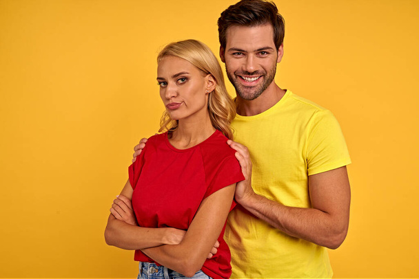 Offended smiling young couple friends in colored t-shirts posing isolated on yellow background studio portrait. People lifestyle concept. Mock up copy space. Holding hands crossed, hugging - Photo, Image