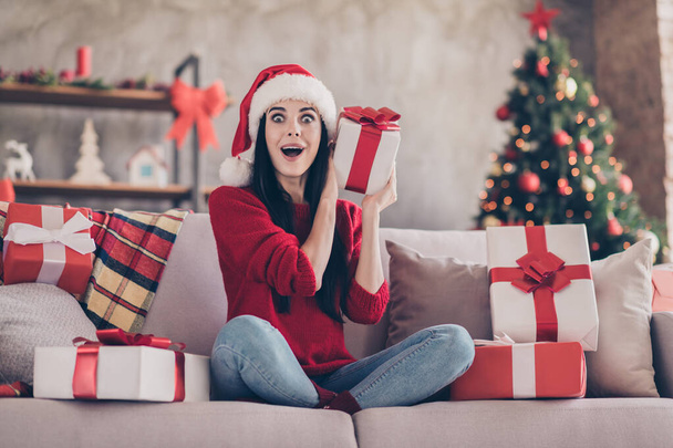 Photo of pretty girl sit surprised face open mouth sofa hold giftbox wear santa cap red sweater jeans in decorated living room indoors - Foto, Bild
