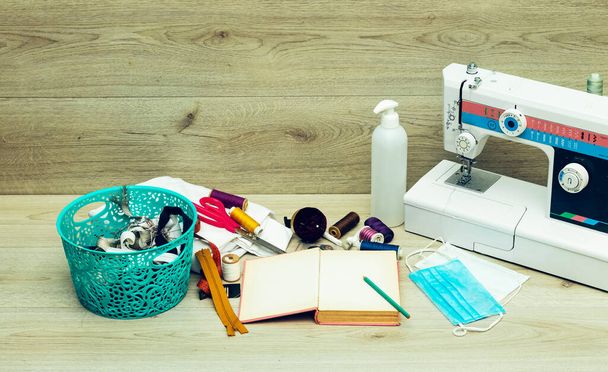 Designer work place sewing machine and medical mask, gel iand  basket of sewing tools : scissors, colorful bobbins with thread and presser foot on gray wooden table - Foto, Bild