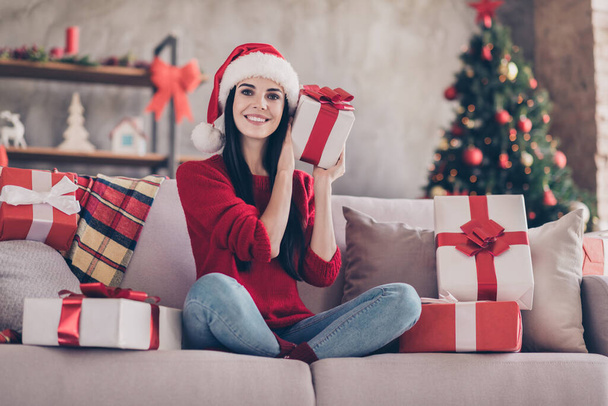 Photo of charming girl sit couch hold present guess wear santa headwear red sweater jeans in decorated living room indoors - Fotó, kép