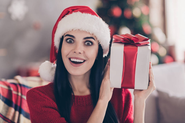 Photo de jolie jeune femme tenir cadeau arc impressionné visage porter Père Noël casque pull rouge dans le salon décoré à l'intérieur - Photo, image