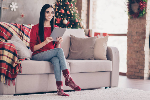Photo of cute young lady sit couch hold tablet toothy smiling wear red sweater denim jeans socks in decorated living room indoors - Foto, afbeelding