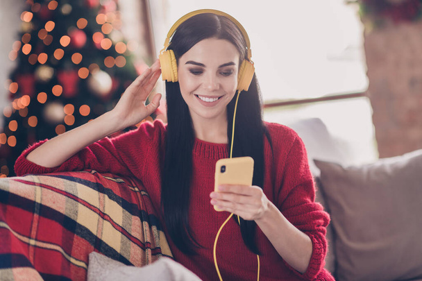 Photo of lovely young girl hold telephone look screen wear headphones red sweater in decorated living room indoors - Photo, Image