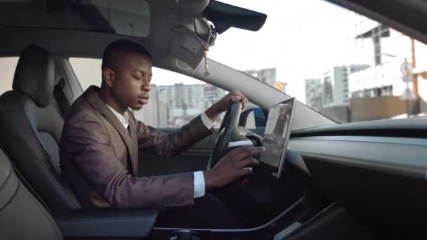 Vista lateral. Joven hombre de negocios negro con café en sus manos sentado en un coche moderno. Dirige su mirada a la cámara. Hombre de negocios exitoso en un coche eléctrico - Imágenes, Vídeo