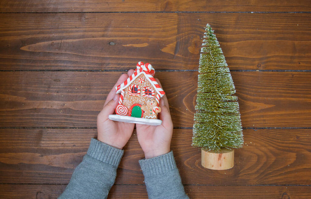 Пряничный домик в детских руках и маленькая елка на деревянном фоне - Фото, изображение
