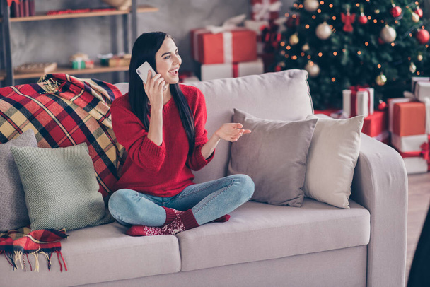 Photo of charming young lady sit sofa hold telephone speak wear red sweater denim jeans socks in decorated living room indoors - Zdjęcie, obraz